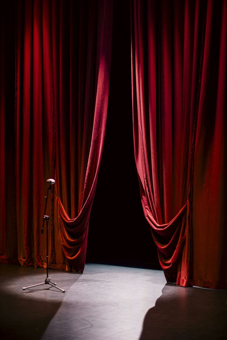 A theater stage with closed red curtains and a single microphone in the spotlight, creating a dramatic ambiance.